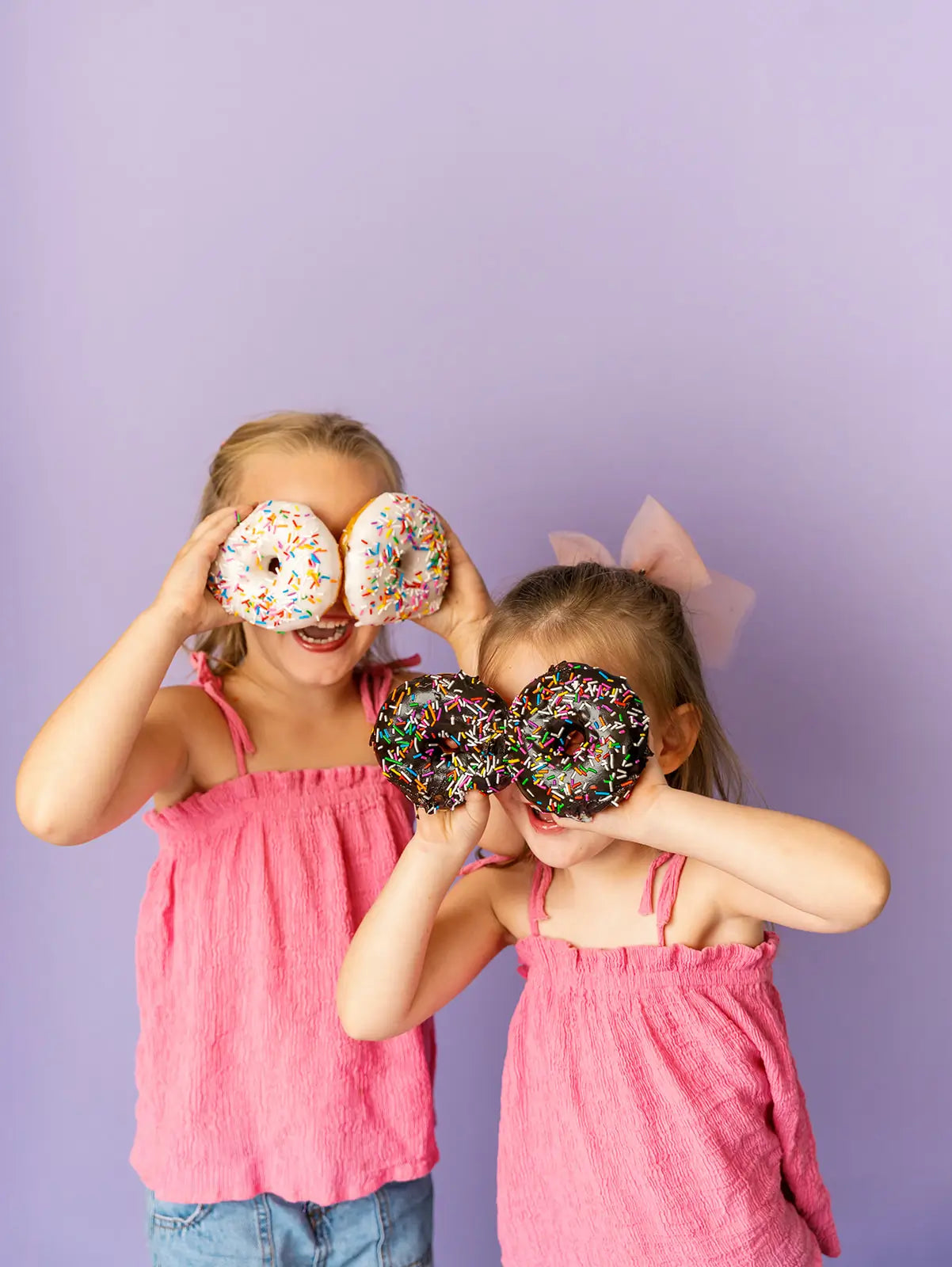Two children in pink tops holding donuts with sprinkles over their eyes against a purple background