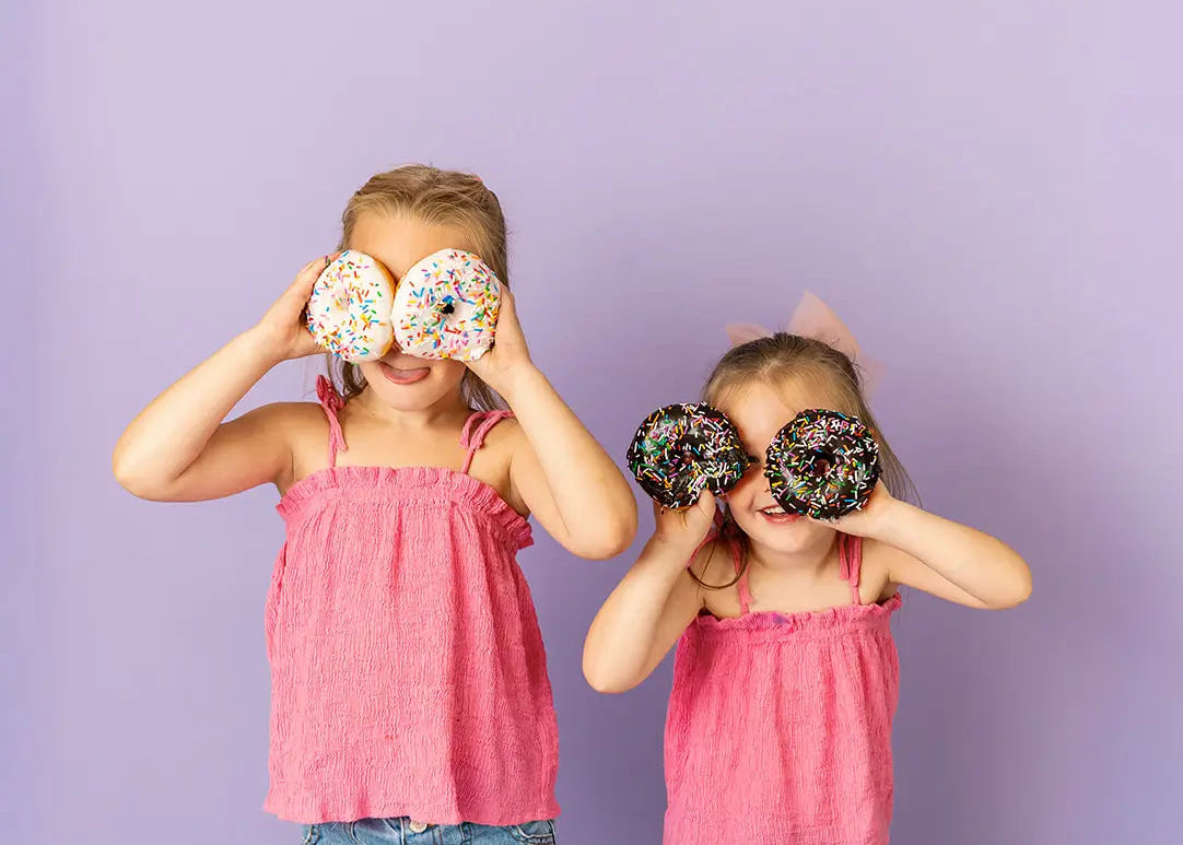 Two children wearing pink tops and jeans, standing against a purple background with confetti on the floor.